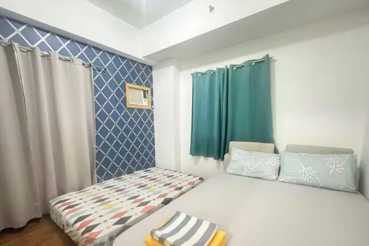 Une chambre avec deux lits, une fenêtre ornée d’un rideau à motifs bleus, un climatiseur fixé au mur et un petit cadre photo.