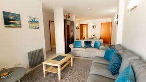 Una sala de estar moderna con un sofá gris, una mesa de centro de madera y una alfombra gris. Hay dos cuadros en la pared, un radiador y una puerta que da a otra habitación.