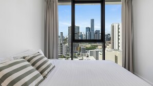 A bedroom with a large bed, a view of a cityscape through a window, and curtains on the window. - 3-Bed City View Apt with Free Parking (South Brisbane)