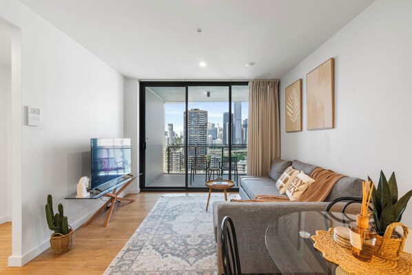 A modern living room with a glass coffee table, a sofa, a TV, and a view of the cityscape through large windows.