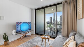 A modern living room with a flat-screen TV, a sofa, a small round table, a cactus plant, and a view of a cityscape through a large sliding glass door. - 3-Bed City View Apt with Free Parking (South Brisbane)