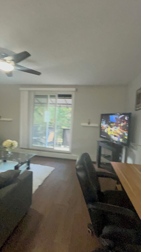 Ein Wohnzimmer mit Fernseher, Deckenventilator, Fenster und Tür zum Ausgang.
