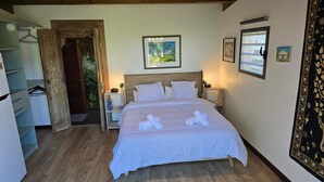 A neatly arranged bedroom with a wooden bed, a small bedside table, a nightstand, a wardrobe, and a door leading to a balcony.