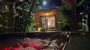 A hot tub with water jets, surrounded by lush greenery and a wooden structure with a glass door.