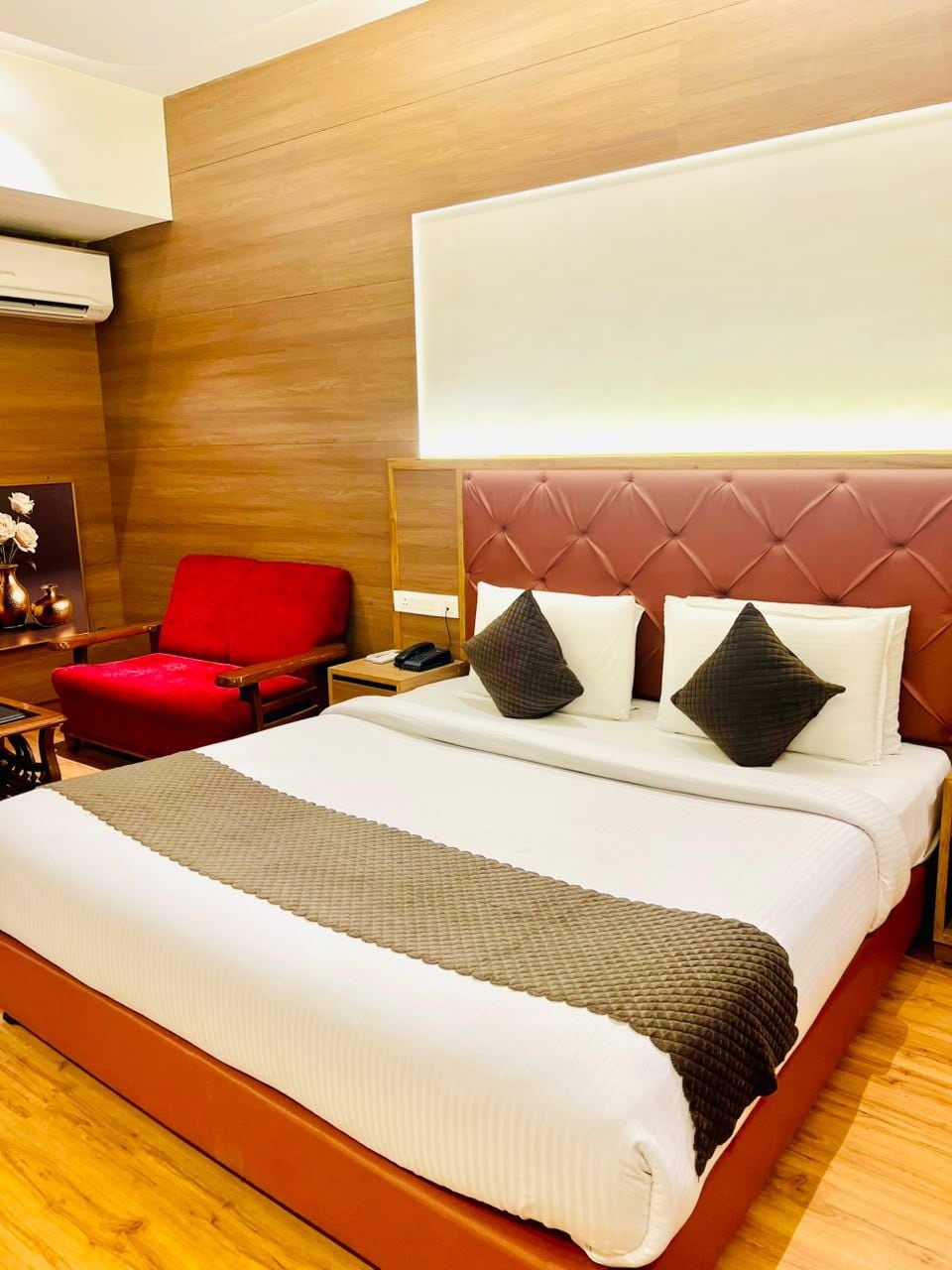 Habitación de hotel con una cama grande, un sillón rojo, una mesita de noche con lámpara y un aire acondicionado instalado en la pared.