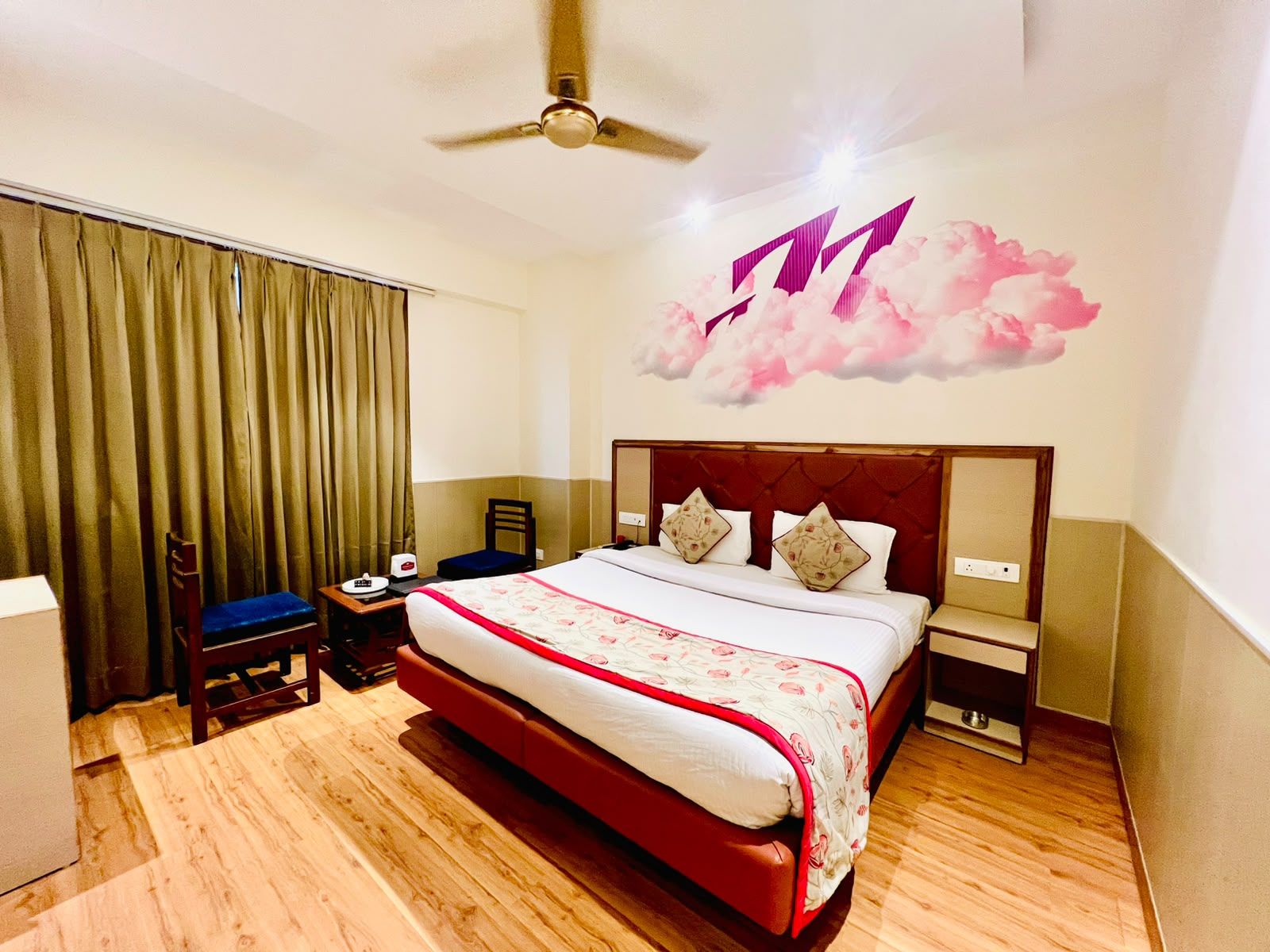 A hotel room with a large bed, a ceiling fan, a desk with a chair, and a wall with a cloud mural.