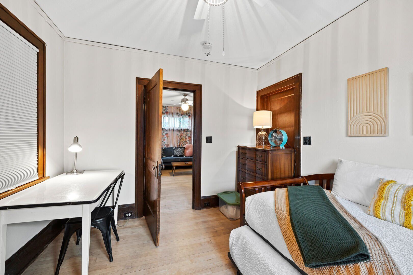 Een hotelkamer met een houten bureau, een bed met een groene plaid, een nachtkastje met een lamp en uitzicht op de aangrenzende kamer.