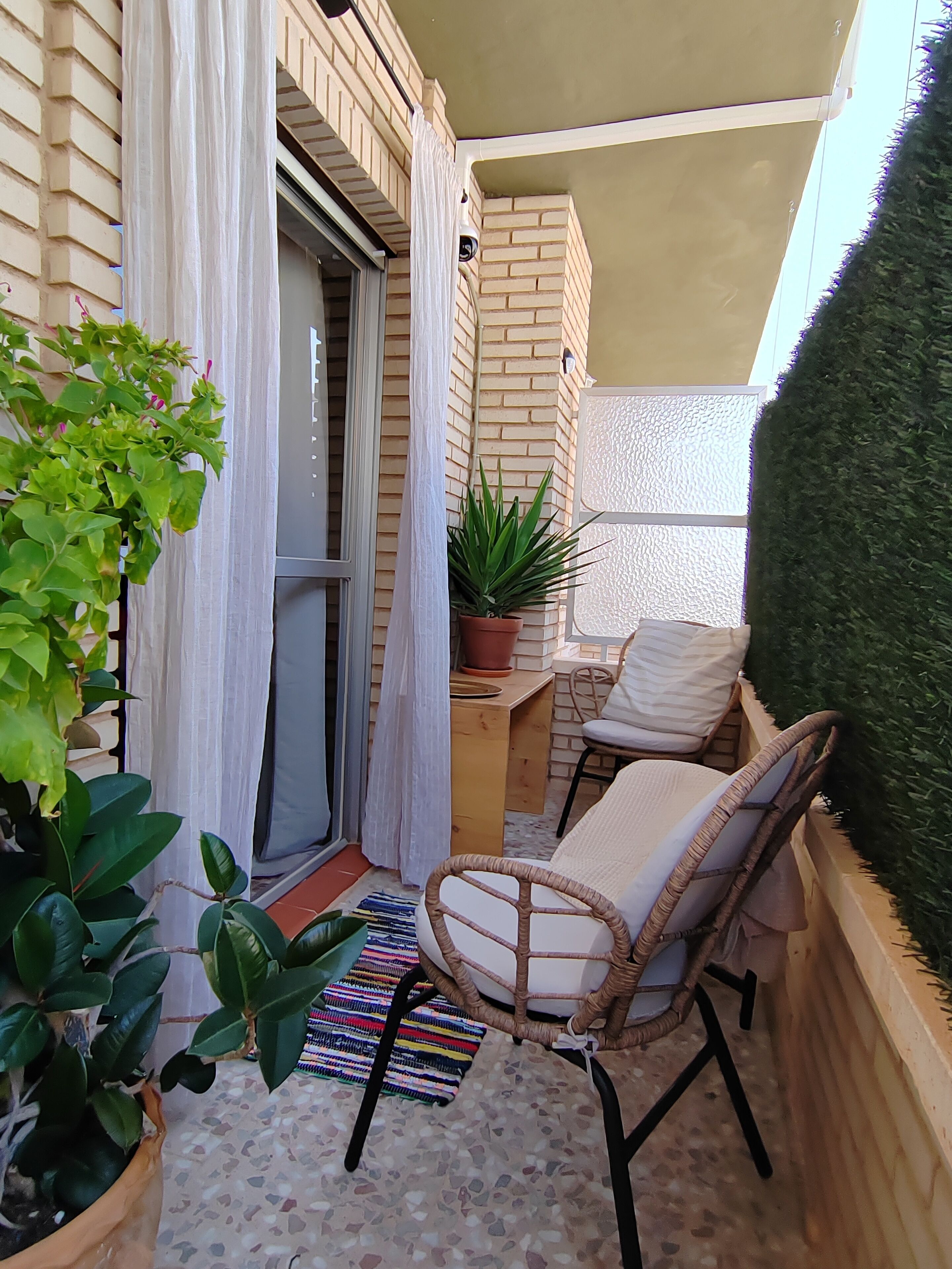 A small balcony with a wicker chair, a potted plant, a small table, and a sliding glass door.