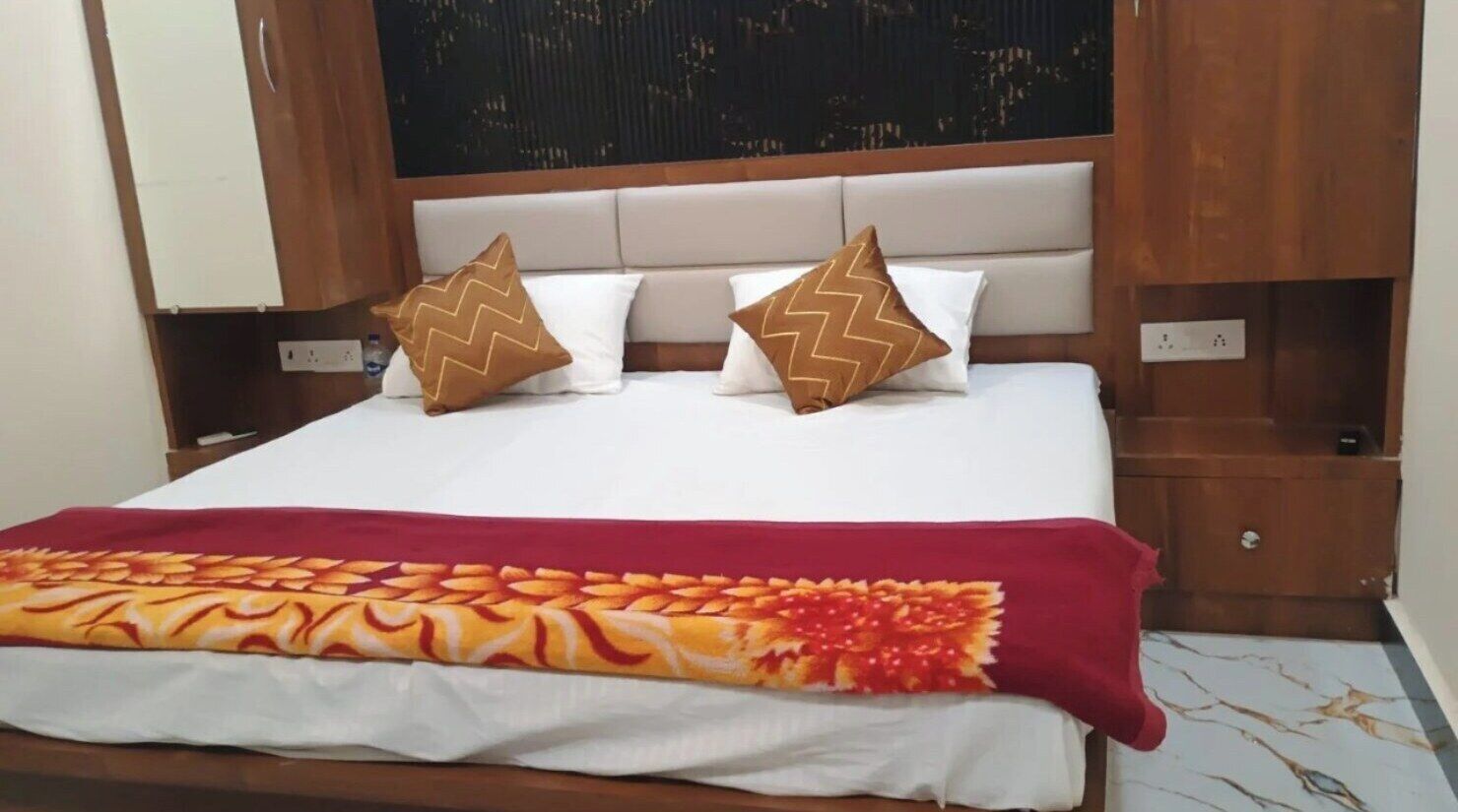 A hotel room with a bed, two pillows, a red and orange blanket, and a wooden headboard.