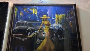Un tableau encadré représentant une femme en robe jaune, debout à côté d’une voiture ancienne, avec une vue sur la ville et une horloge en arrière-plan.