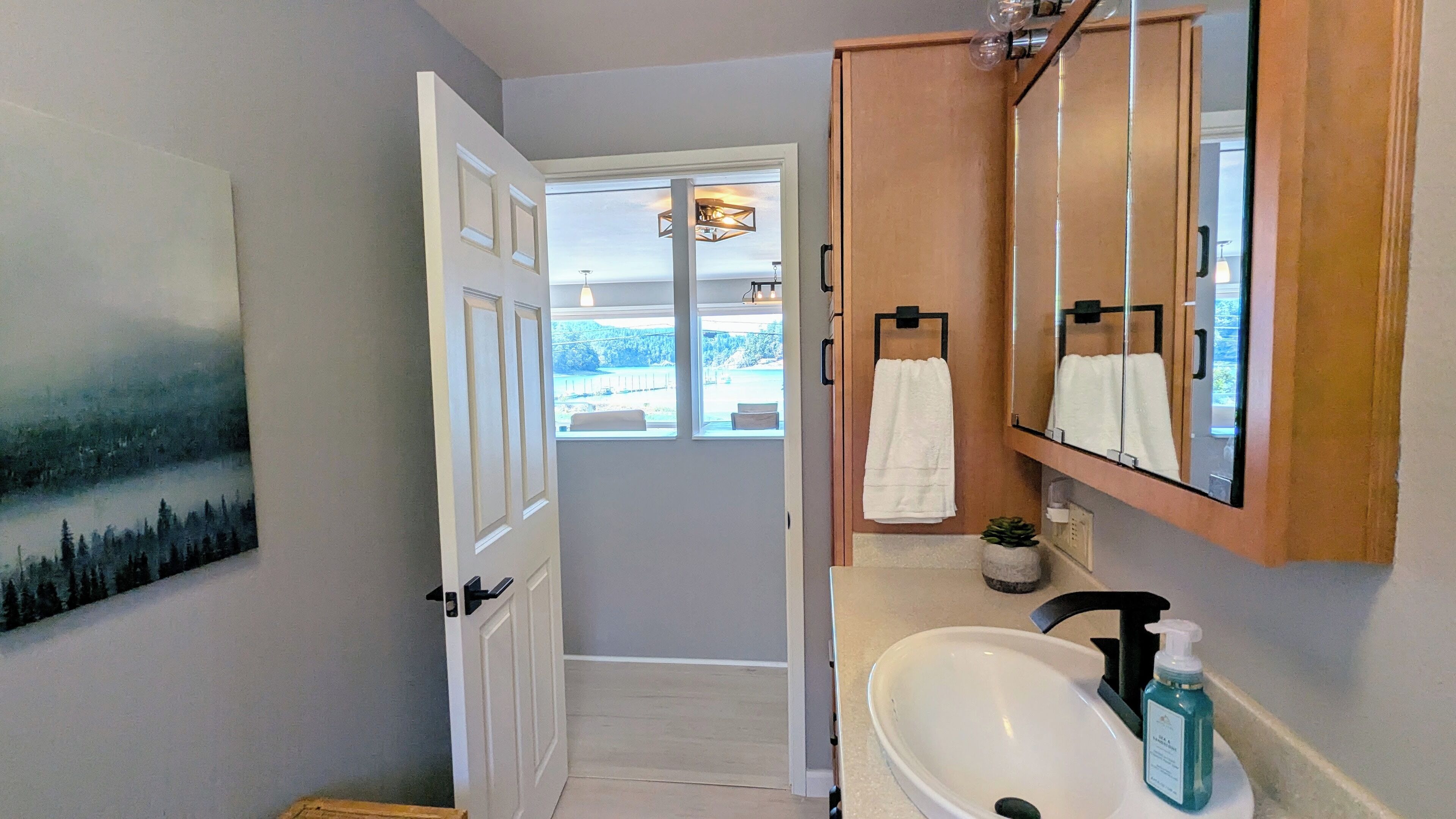 A bathroom with a white sink, a mirror, and a wooden cabinet. There is a painting on the wall and a window with a view of the outdoors.