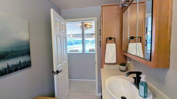 A bathroom with a white sink, a mirror, and a wooden cabinet. There is a painting on the wall and a window with a view of the outdoors.