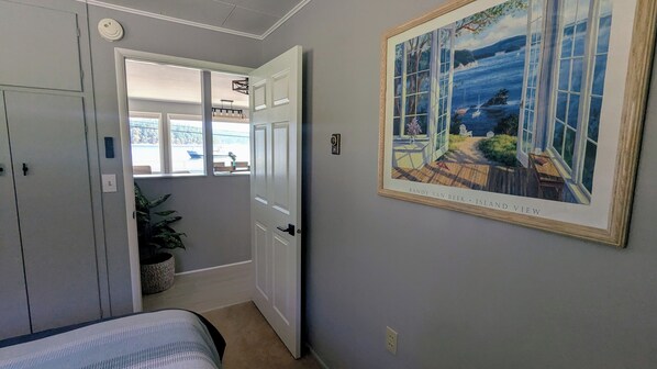 A bedroom with a bed, a door leading to a balcony, a potted plant, and a painting of an island view.