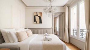 A neatly arranged bedroom with a bed, a chandelier, a painting, and a view through a window. - Trocadero - Elegant apartment with lift (Unknown)
