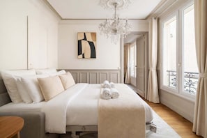 A neatly arranged bedroom with a bed, a chandelier, a painting, and a view through a window.