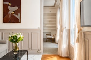 Quarto com sofá, mesa de centro com um vaso de flores, pintura na parede, televisão e janela com cortinas.