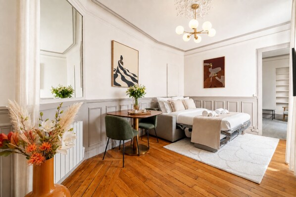 A hotel room with a bed, a desk, a chair, a vase with flowers, and artwork on the walls.