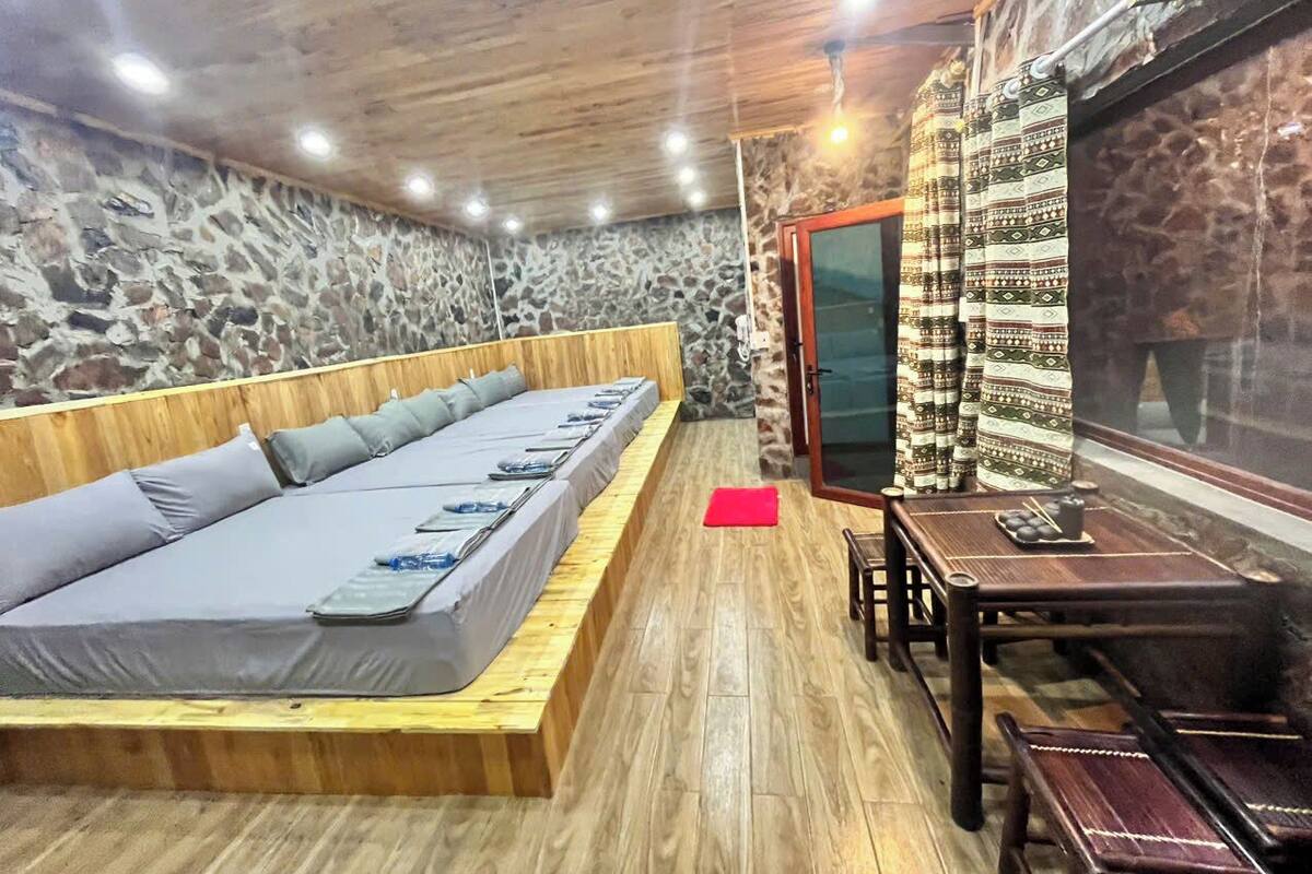 A room with a long wooden bed, a small table with a tray, and a stone wall.