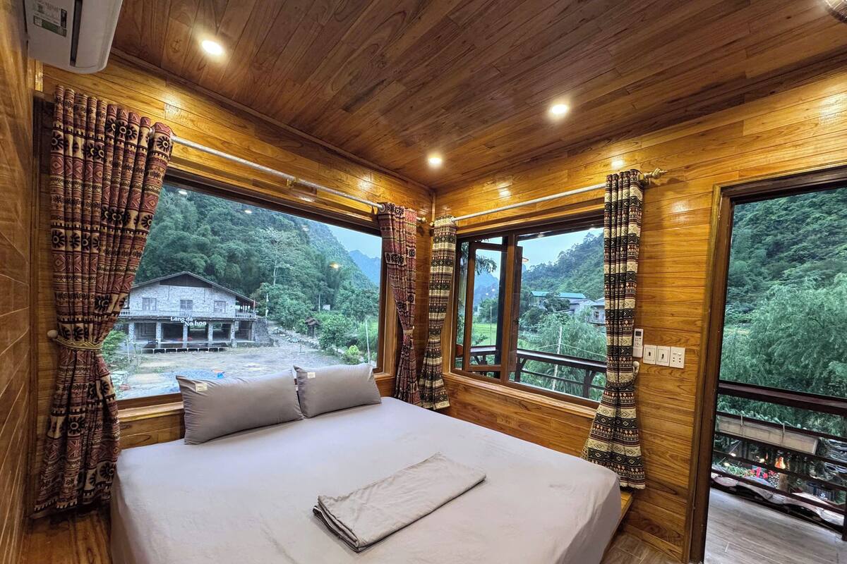 A wooden cabin room with a bed, large windows, and a view of a mountain and a building outside.