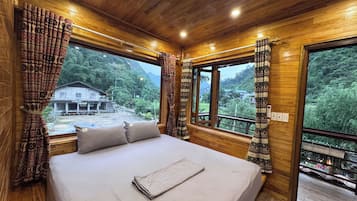 A wooden cabin room with a bed, large windows, and a view of a mountain and a building outside.