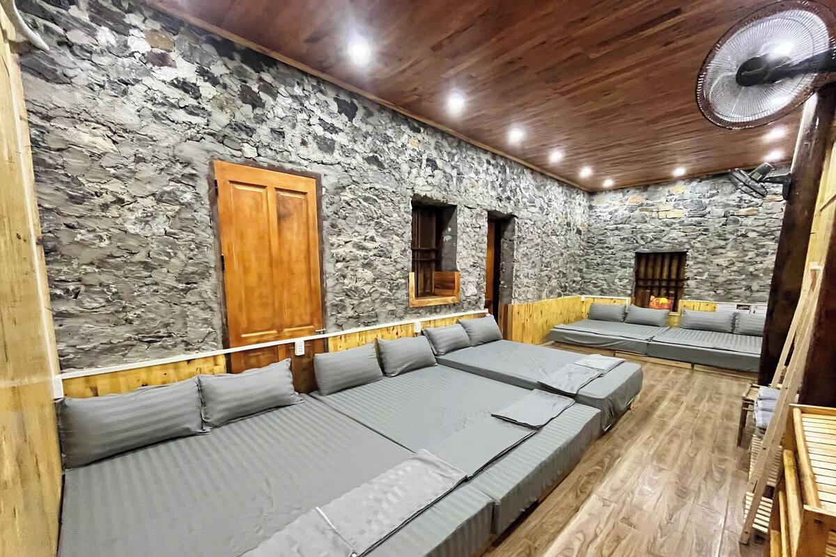 A room with a stone wall, wooden ceiling, and a large fan. There are two beds with grey bedding and wooden headboards.