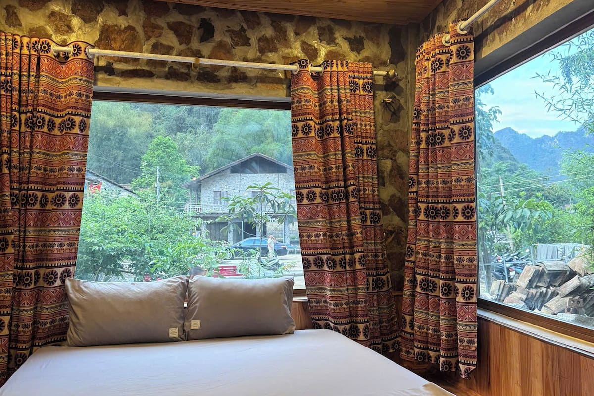 A bedroom with a wooden ceiling, a bed with a white mattress and two pillows, patterned curtains, and a window with a view of greenery and a building.