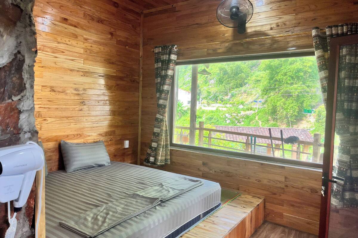 A wooden cabin room with a bed, a ceiling fan, and a window with curtains.