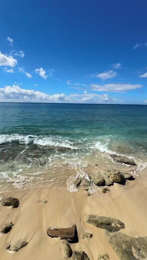 Une plage à l’eau d’un bleu limpide, avec un rivage sablonneux et quelques rochers épars.