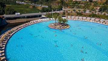 A large outdoor swimming pool with multiple lanes, surrounded by lounge chairs and umbrellas, set against a backdrop of hills and a parking lot.