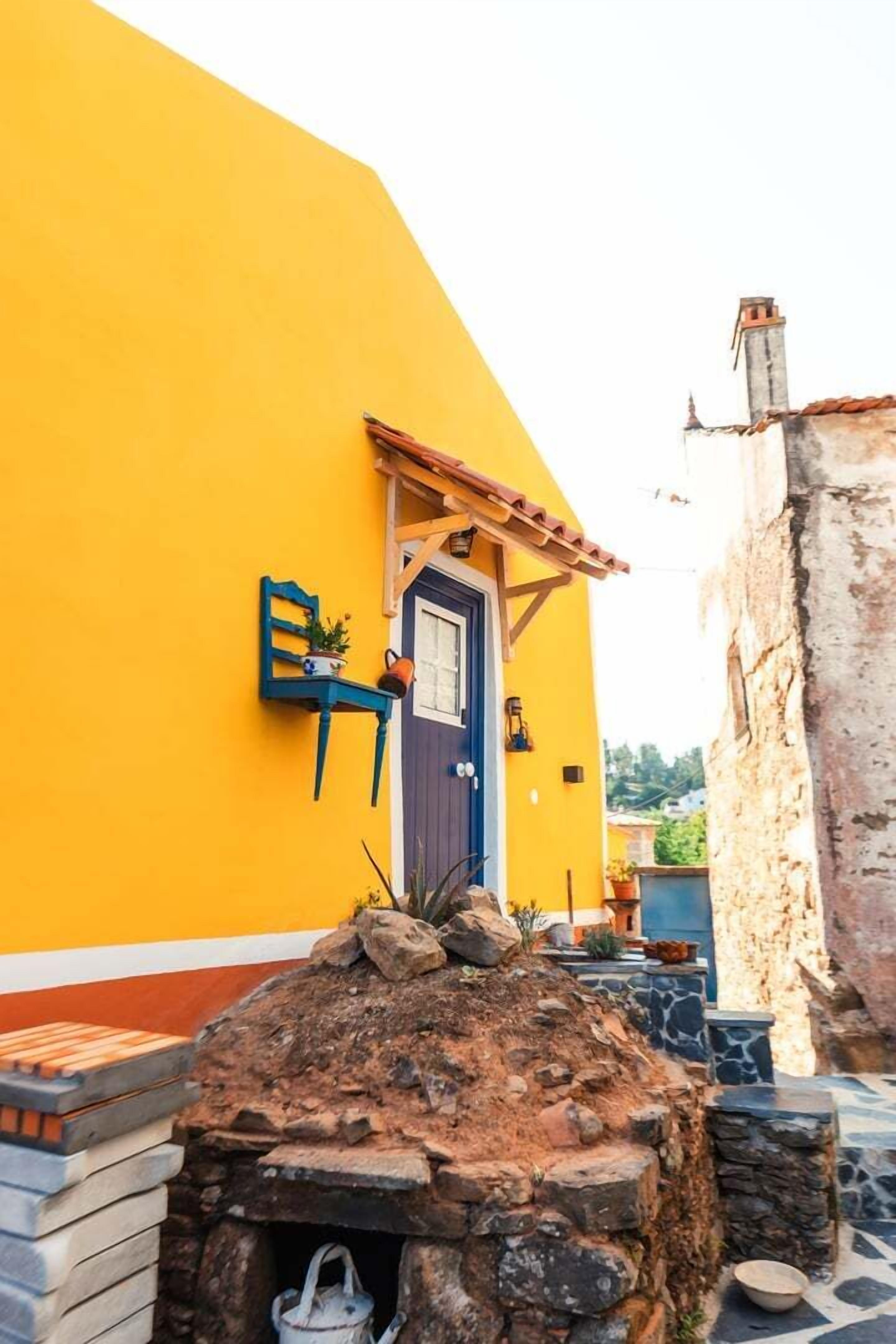 A yellow building with a blue door, a stone oven, and a small garden area.