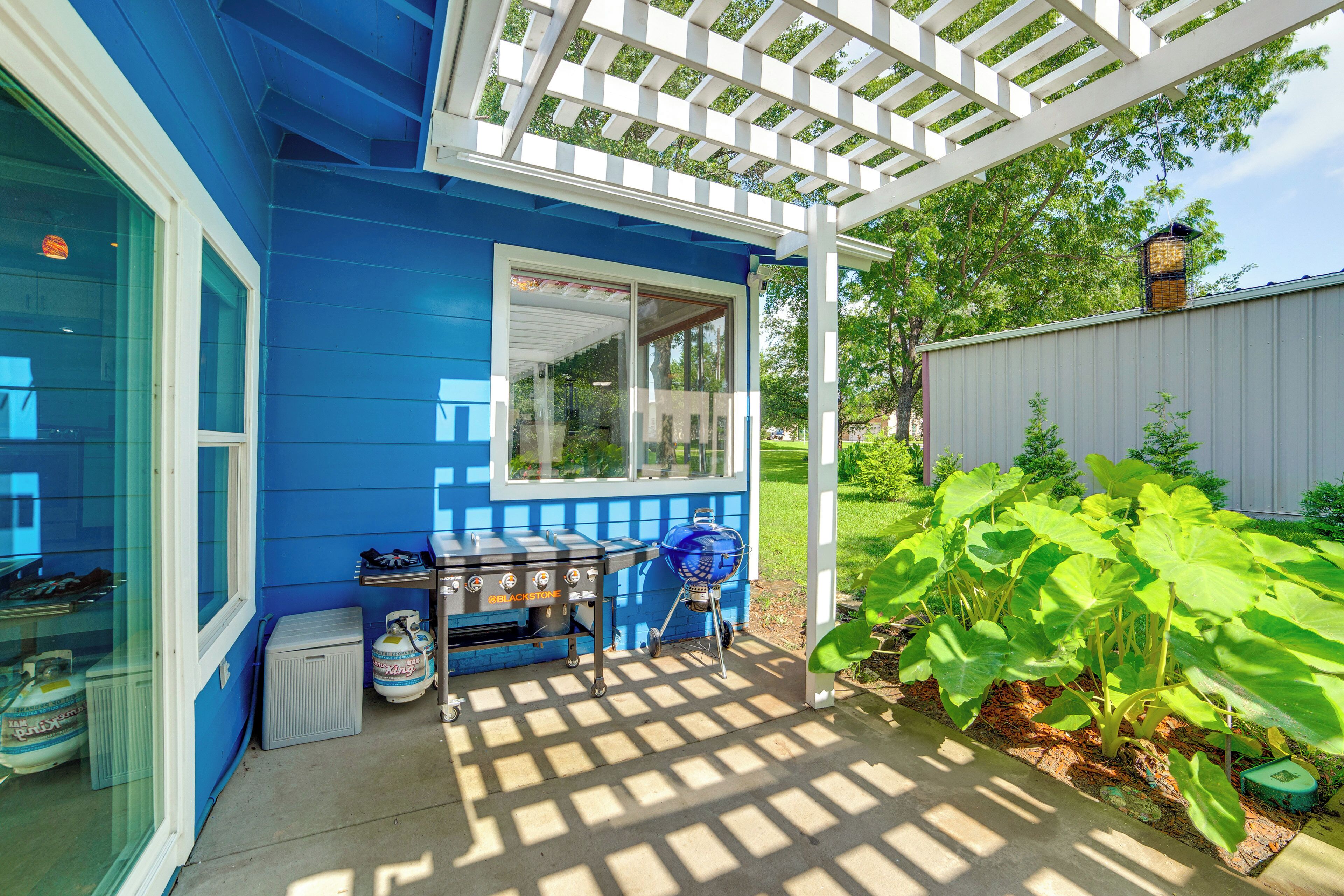 Une maison bleue avec un patio couvert, un barbecue et un jardin où poussent de grandes plantes à feuilles larges.