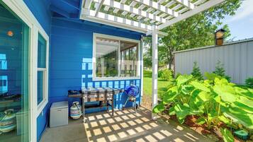 Une maison bleue avec un patio couvert, un barbecue et un jardin où poussent de grandes plantes à feuilles larges.