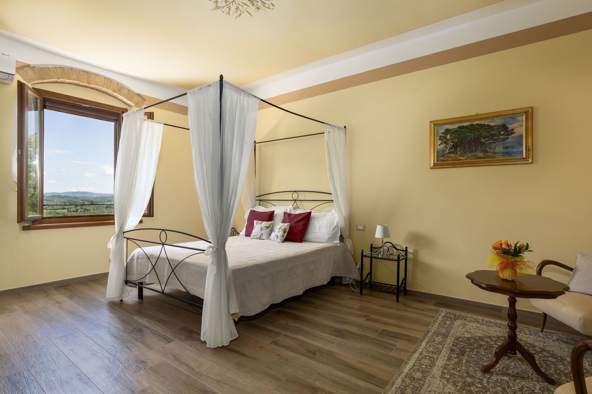 A bedroom with a four-poster bed, a view through a window, a painting on the wall, and a small table with a vase.