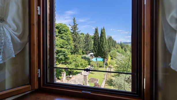 Vista desde una ventana con cortinas, que da a un jardín cuidado con piscina, construcciones de piedra y altos árboles.