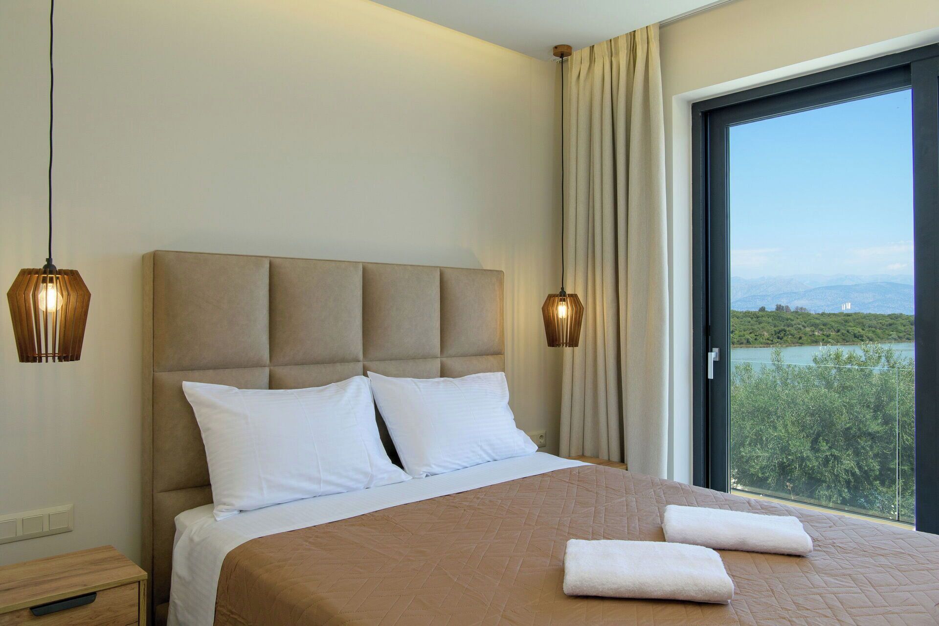 A hotel room with a large bed, two bedside lamps, a nightstand, and a view of a lake and mountains.