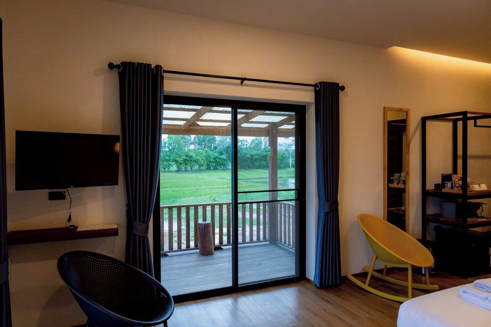 Una habitación de hotel con televisión, una silla, una cama, un balcón con cortinas y vistas a un paisaje verde.