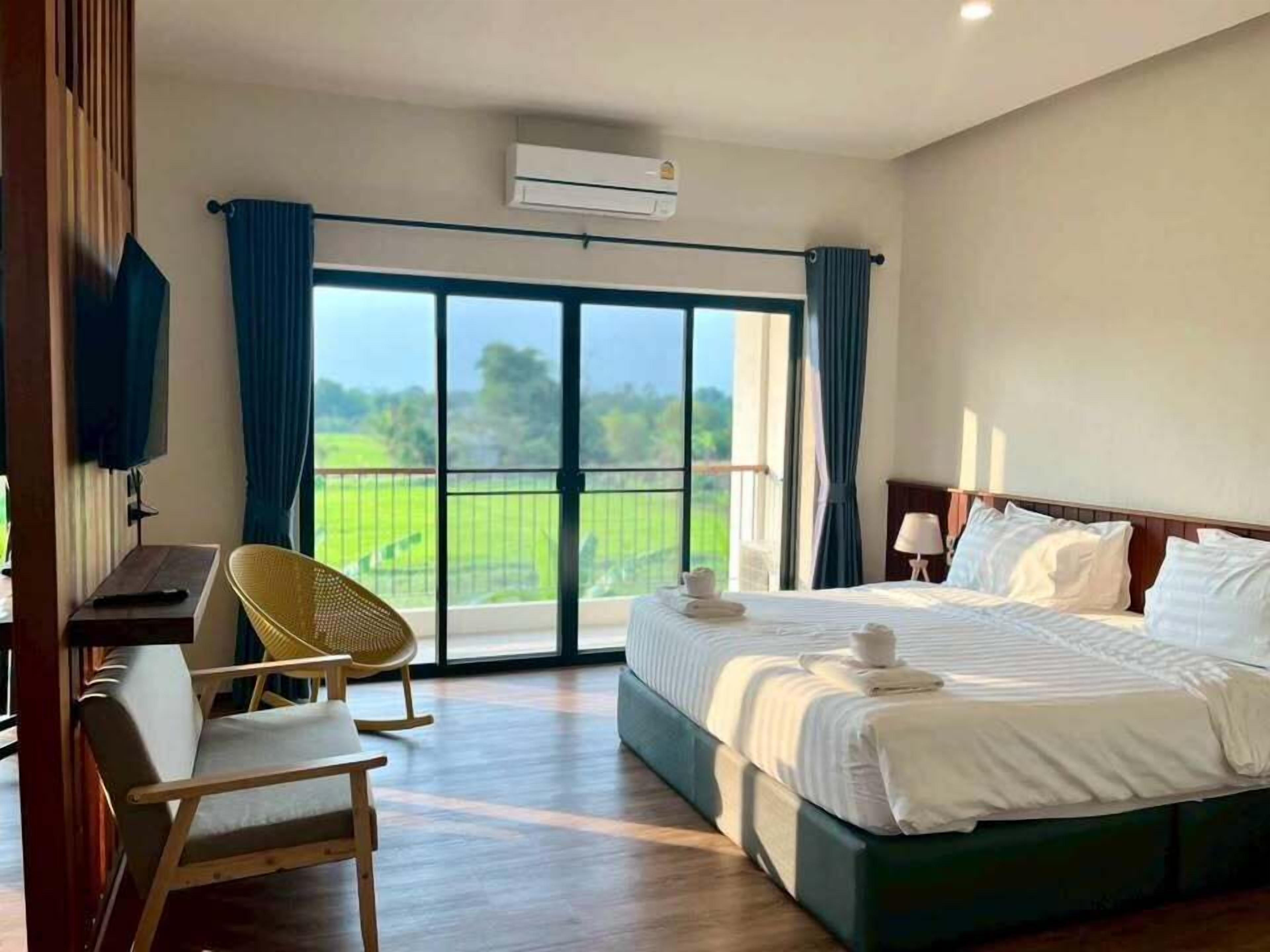 Habitación de hotel con cama, escritorio, silla, equipo de aire acondicionado y vista a un paisaje verde a través de un ventanal grande.