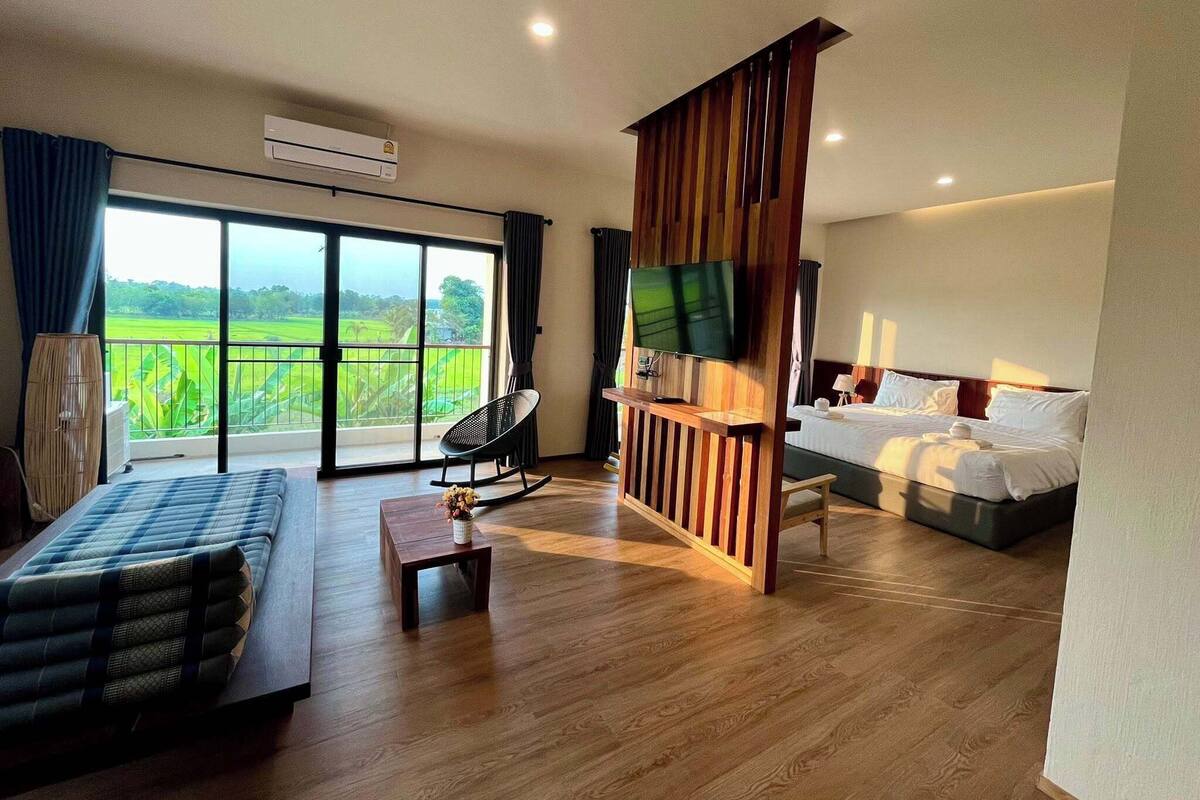 Ein Hotelzimmer mit Bett, Fernseher, Sofa, Balkon mit Blick auf eine grüne Landschaft und einer Holzabtrennung.