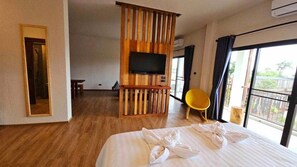 A hotel room with a large bed, a wooden partition, a flat-screen TV, a yellow chair, and a balcony with a view.