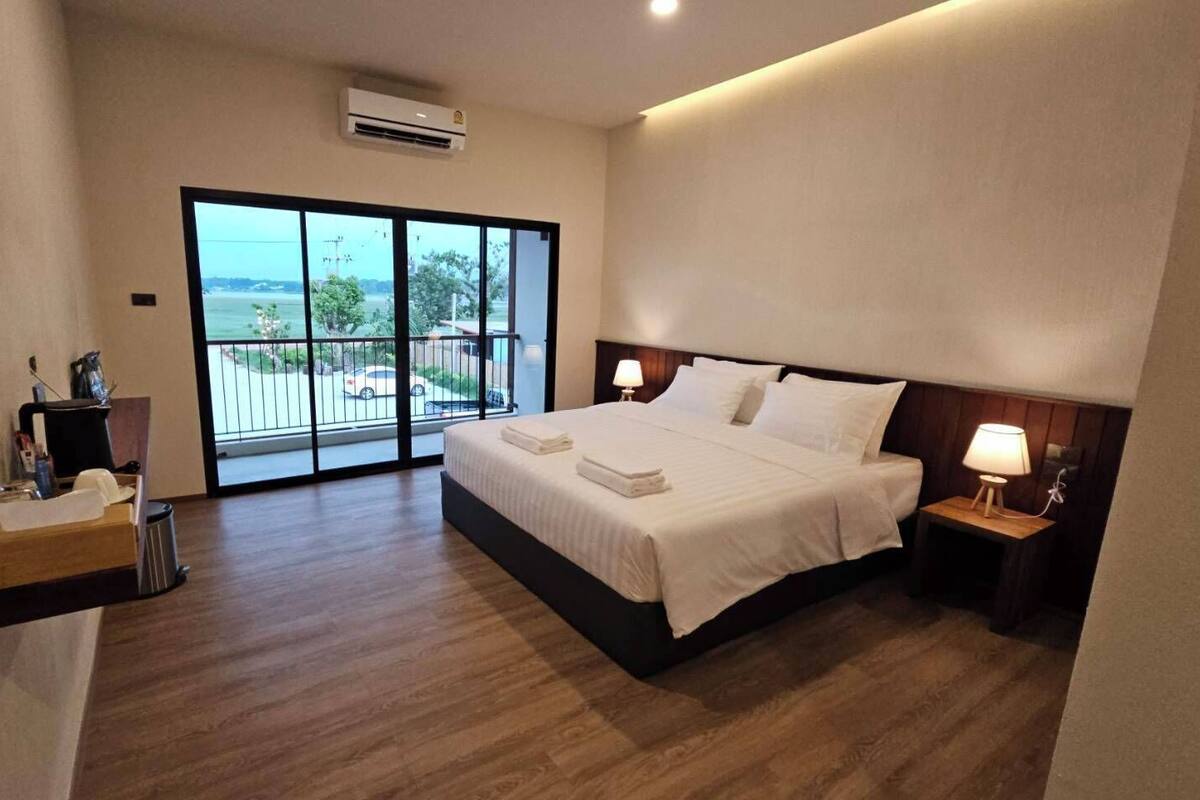Ein Hotelzimmer mit einem großen Bett, einem Nachttisch mit Lampe, einem kleinen Schreibtisch mit Stuhl und Blick durch eine Schiebetür ins Freie.