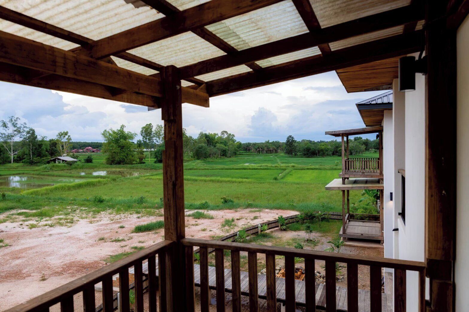 A wooden balcony with a view of a green rice field and a distant red building.