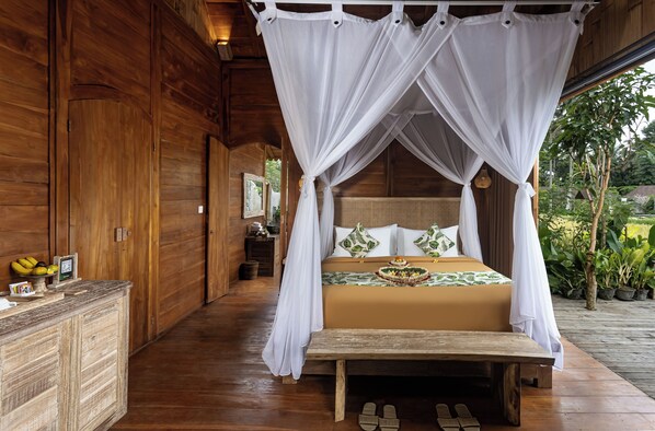 A wooden room with a bed under a canopy, a wooden bench, and a small table with a picture frame and a clock.