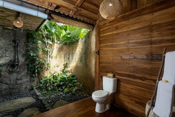 Un baño con detalles de madera, un inodoro, un lavamanos y una pequeña zona ajardinada con plantas y un sendero de piedra.
