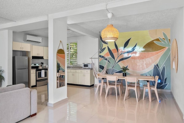A modern kitchen with white cabinets, a stainless steel refrigerator, and a microwave. There is a dining area with a wooden table and chairs, and a large abstract painting on the wall.