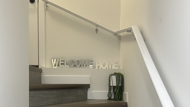 A staircase with a handrail, a welcome home sign, and a small plant on the landing. - Cozy Corner Rentals (Saskatoon)