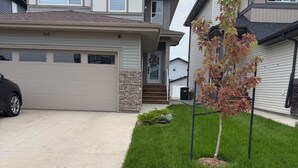 A two-story house with a garage, a driveway, and a well-maintained lawn with a small tree. - Cozy Corner Rentals (Saskatoon)