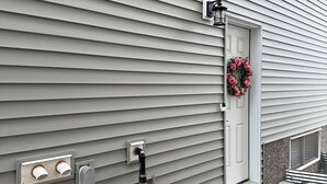 A house with a grey siding exterior, a white door with a pink wreath, and a small porch with a concrete step. - Cozy Corner Rentals (Saskatoon)