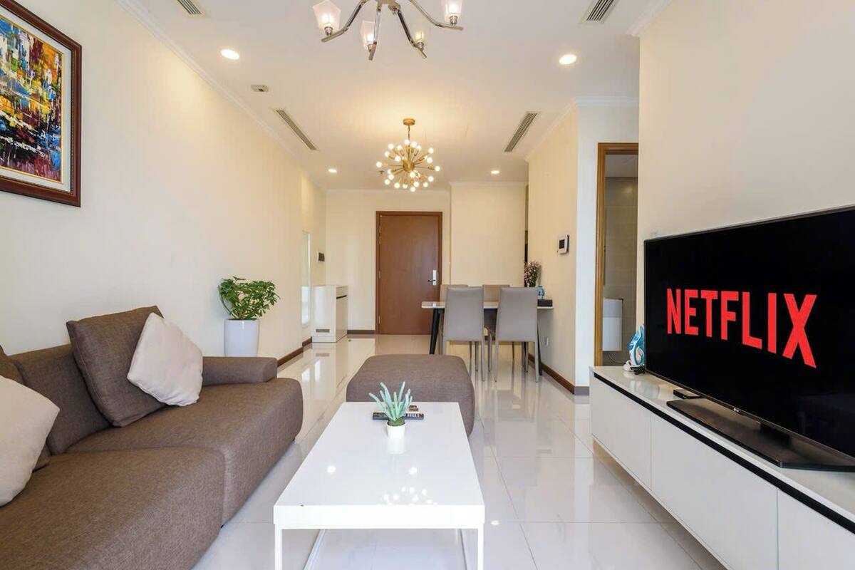 A modern living room with a sofa, a coffee table, a TV displaying 'NETFLIX', and a dining area in the background.