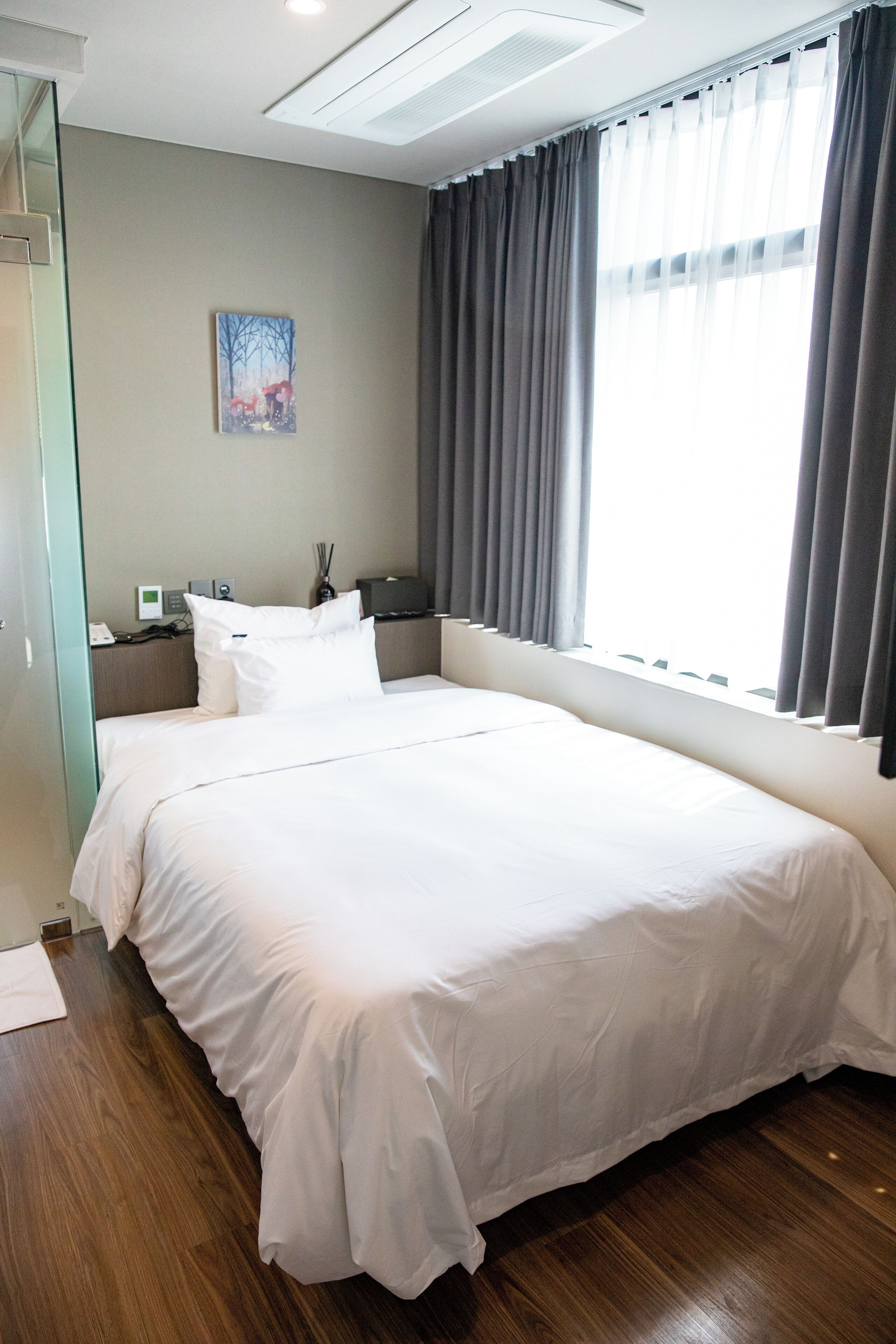 A hotel room with a large bed, a bedside table with a lamp, a painting on the wall, and a window with curtains.