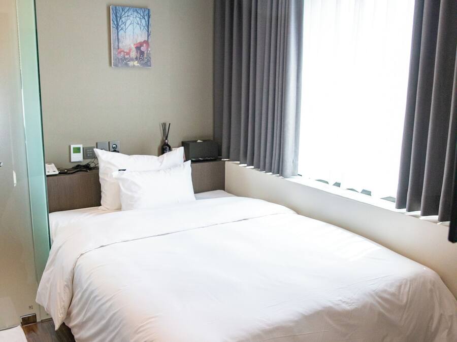 A hotel room with a large bed, a bedside table with a lamp, a painting on the wall, and a window with curtains.